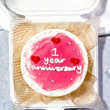 A pink cake with '1 year anniversary' written on it, with small red hearts and white dots, in a box