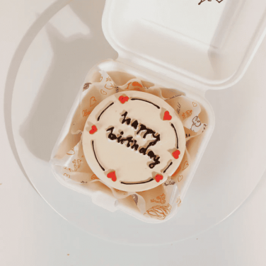 A small white birthday cake with 'Happy Birthday' written in chocolate, decorated with tiny red heart-shaped candies, inside a white container.