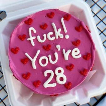 A bold pink cake with a humorous age declaration in white icing, surrounded by small red heart decorations, presented in a white box.