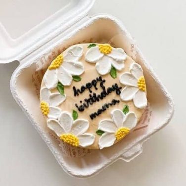 A round cake with white icing, decorated with white and yellow daisies, and 'Happy Birthday mum' written in black icing,