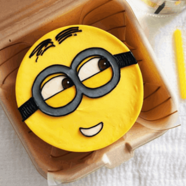 A yellow round cake designed to look like a minion's face with black-rimmed goggles, smiling, placed in a clear bento box.
