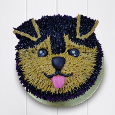 Round dog birthday cake designed like a cute puppy face with black and tan fur, pink tongue, and shiny eyes