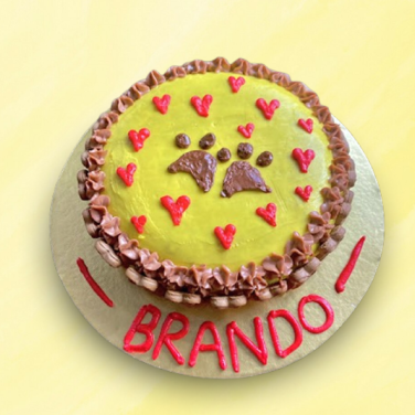 Round yellow dog cake with brown paw prints and red hearts, decorated with chocolate frosting and personalized name